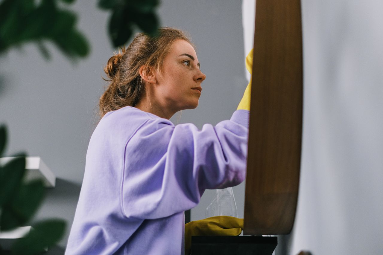 Vue latérale d’une jeune femme concentrée, portant des gants en latex, debout et nettoyant un miroir dans une pièce lumineuse en journée